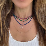 A close-up of a person's neck and upper chest wearing four layered necklaces: a thin gold chain, a blue lapiz beaded chain with a silver diamond horseshoe charm, a green malachite beaded chain with a small diamond heart charm, and a red beaded chain ruby