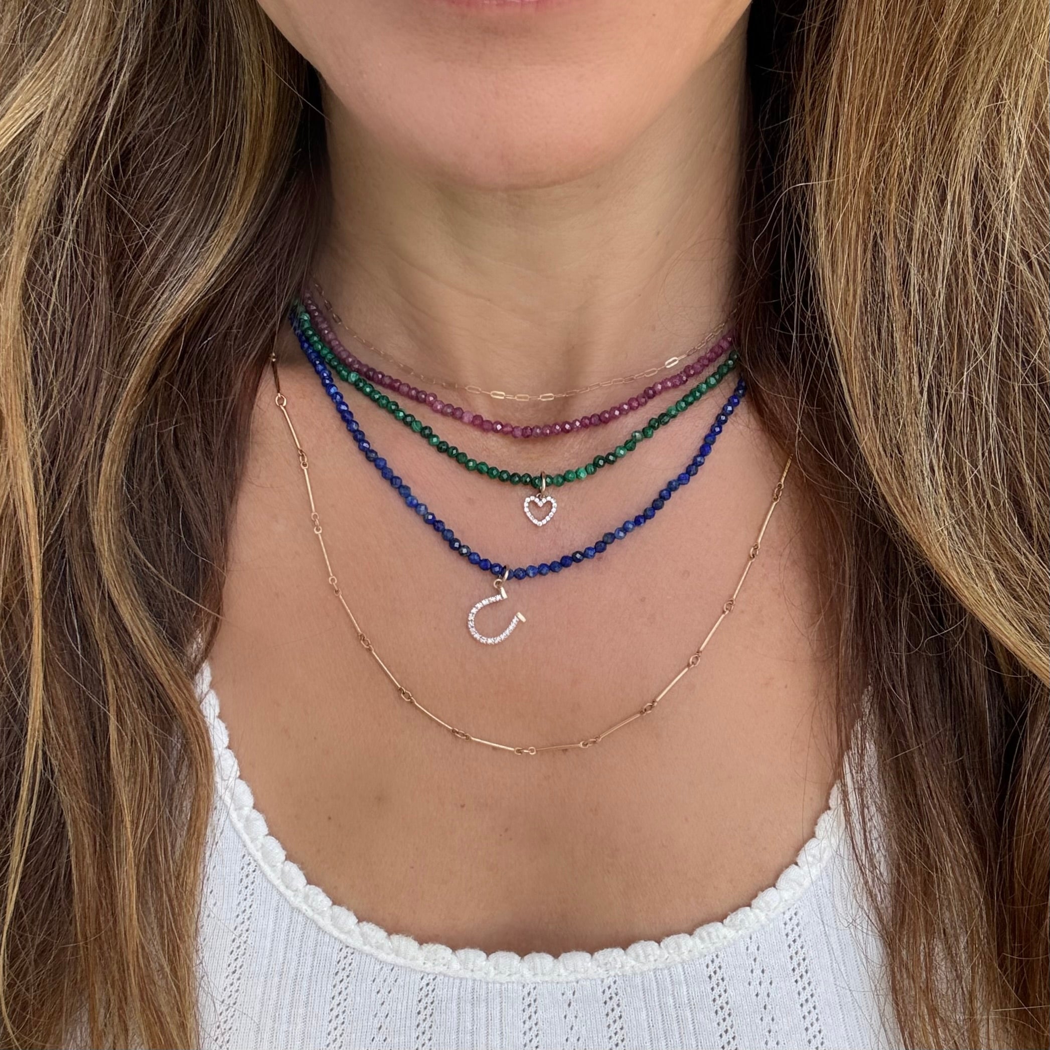 A close-up of a person's neck and upper chest wearing four layered necklaces: a thin gold chain, a blue lapiz beaded chain with a silver diamond horseshoe charm, a green malachite beaded chain with a small diamond heart charm, and a red beaded chain ruby