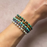 A close-up of a person's wrist wearing a stack of five gemstone beaded stretch bracelets. The bracelets feature a mix of turquoise, malachite, lapis blue, pearl white, and tigers eye brown beads with metallic gold and silver accents