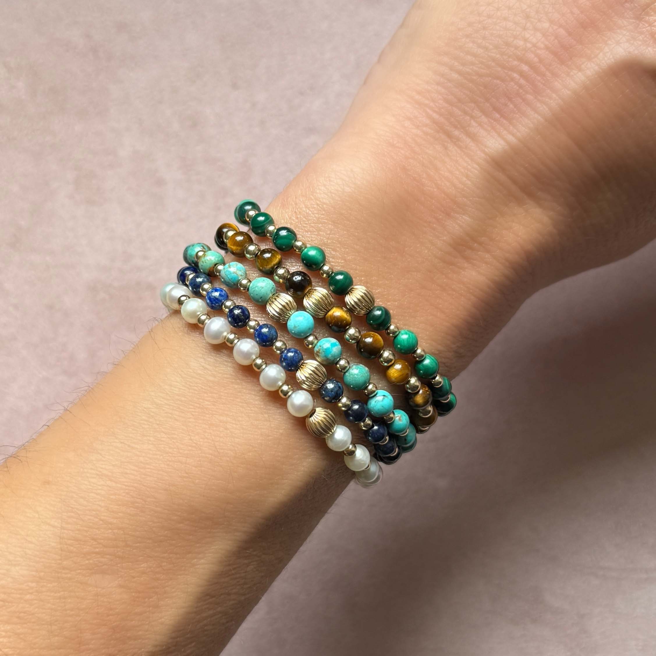 A close-up of a person's wrist wearing a stack of five gemstone beaded stretch bracelets. The bracelets feature a mix of turquoise, malachite, lapis blue, pearl white, and tigers eye brown beads with metallic gold and silver accents