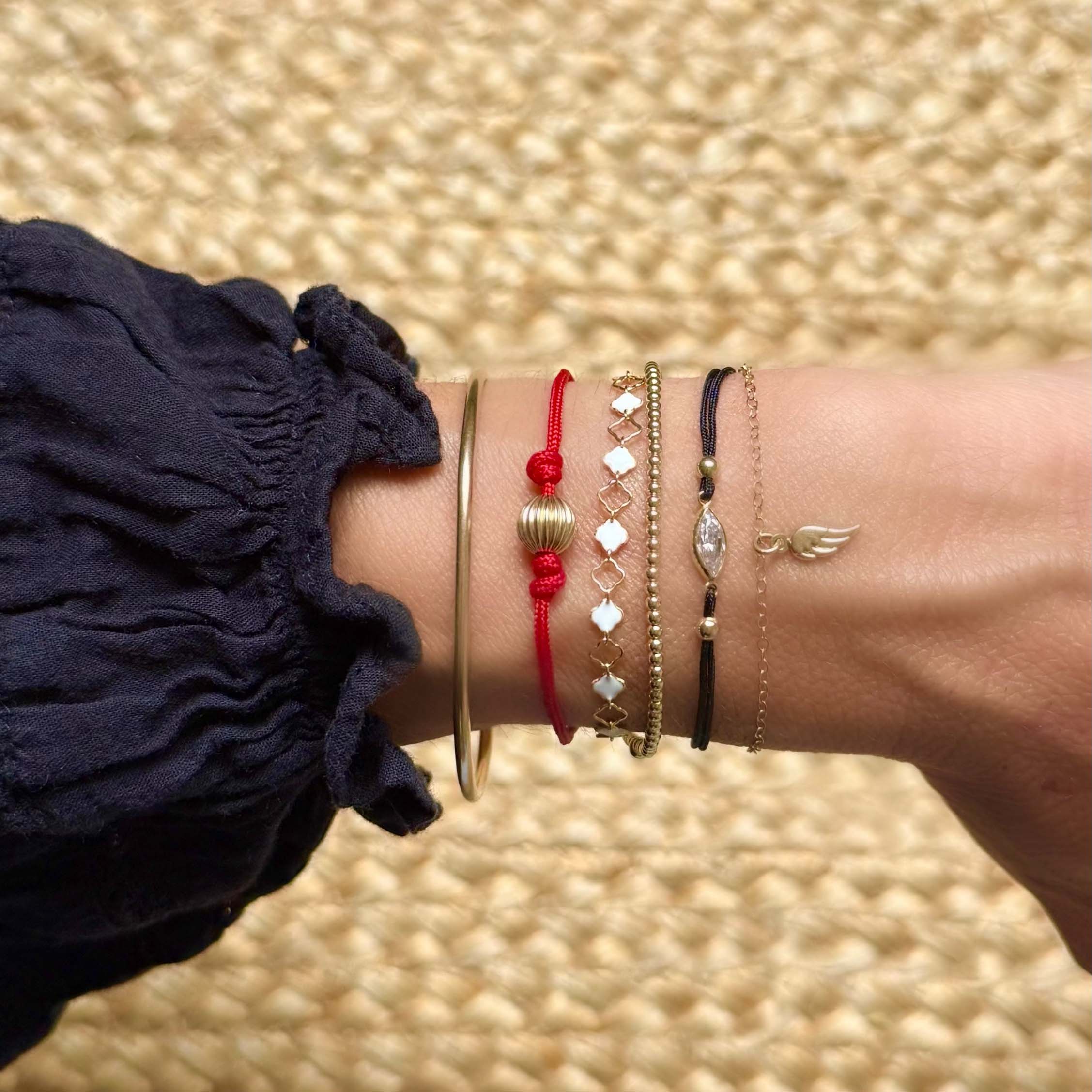 A close-up of a person's wrist wearing a stack of delicate bracelets, including a gold bangle, a red cord bracelet, , a dark beaded bracelet, and a thin gold filled chain with a wing charm. 