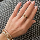 A close-up of a hand wearing a gold crescent moon ring on the ring finger, along with layered gold and silver bracelets, set against a black and white textured woven background.