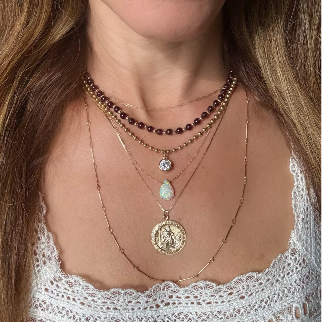 A close-up of a woman wearing a layered necklace stack with multiple gold-tone chains, a garnet beaded strand, a small clear stone pendant, a opal teardrop stone pendant, and a large round gold coin  St Christopher pendant.