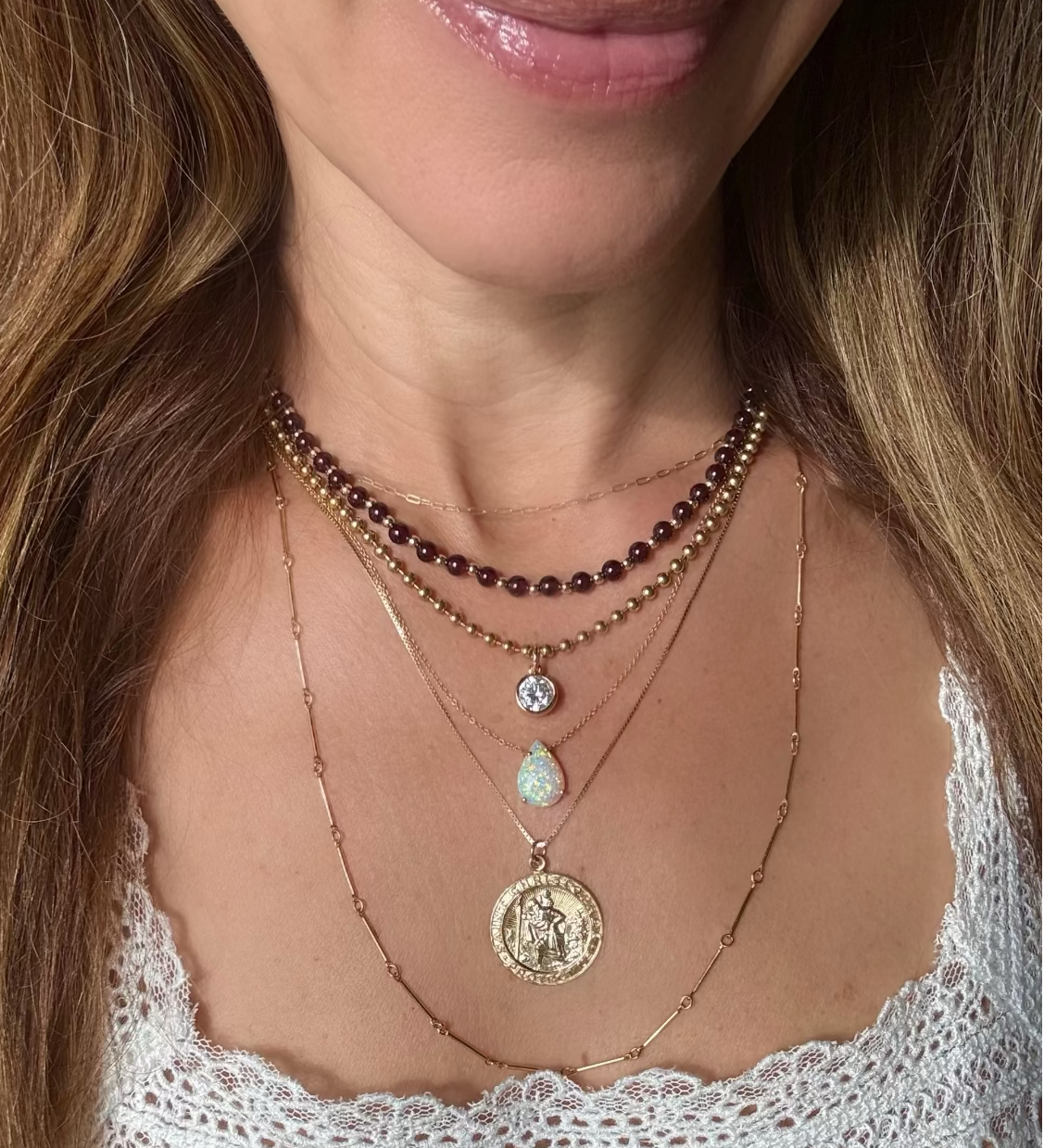 A close-up of a woman wearing a layered necklace stack with multiple gold-tone chains, a garnet beaded strand, a small clear stone pendant, a opal teardrop stone pendant, and a large round gold coin  St Christopher pendant.