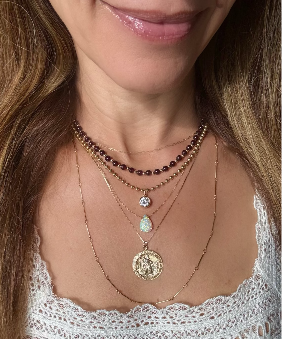A close-up of a woman wearing a layered necklace stack with multiple gold-tone chains, a garnet beaded strand, a small clear stone pendant, a opal teardrop stone pendant, and a large round gold coin  St Christopher pendant.