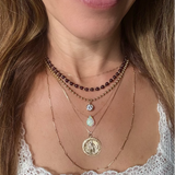A close-up of a woman wearing a layered necklace stack with multiple gold-tone chains, a garnet beaded strand, a small clear stone pendant, a opal teardrop stone pendant, and a large round gold coin  St Christopher pendant.