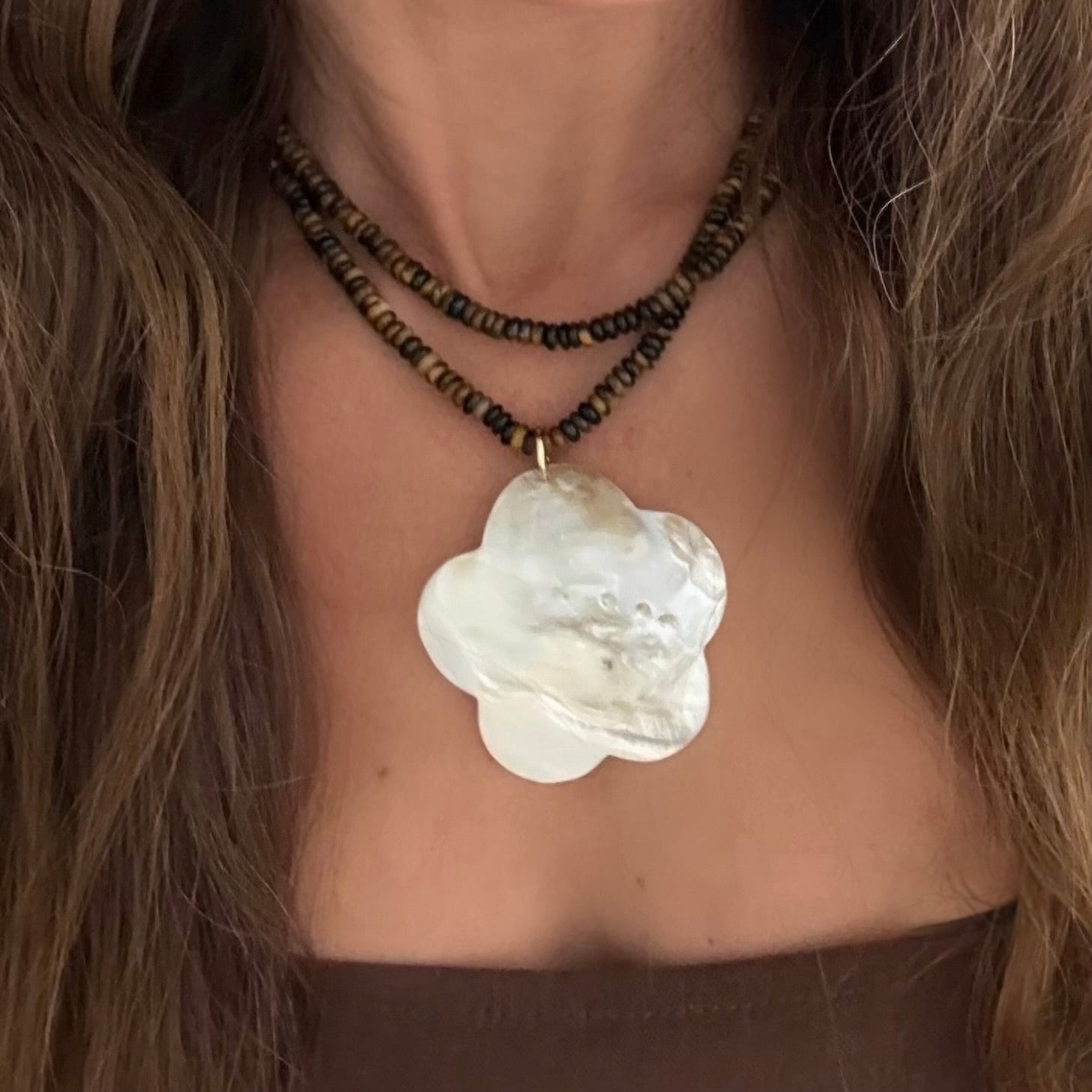 A close-up of a person wearing a double-strand tigers eye bead necklace with dark brown and beige beads and a large, flower-shaped white mother of pearl pendant.